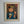 A framed vintage oil painting of a young girl holding a bowl and chopsticks, shown hanging on a light gray wall. The artwork has a warm brown and red background with the girl in a blue garment, enclosed in a light wood frame. Very mid century
