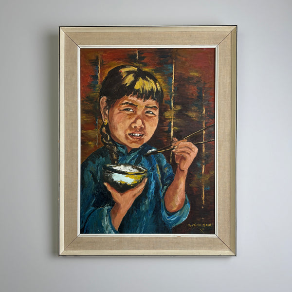 A framed vintage oil painting of a young girl holding a bowl and chopsticks, shown hanging on a light gray wall. The artwork has a warm brown and red background with the girl in a blue garment, enclosed in a light wood frame. Very mid century