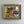 A framed still life mid century oil painting featuring a green coffee pot, a dark cup, and yellow-orange table elements, housed in a textured gold frame against a plain white wall.
