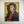 A vintage mid century framed portrait oil painting of Elvis Presley on a white wall. The painting depicts Elvis with dark hair, a black jacket, and a bright red scarf against a textured green background.