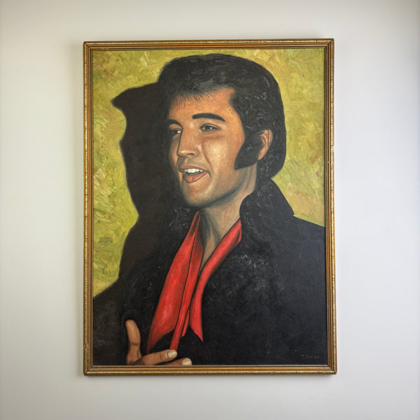 A vintage mid century framed portrait oil painting of Elvis Presley on a white wall. The painting depicts Elvis with dark hair, a black jacket, and a bright red scarf against a textured green background.