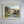 A vintage framed landscape oil painting hanging on a light gray wall. The painting depicts a winding road through rolling hills under a pale sky, enclosed in a distressed off-white wooden frame.