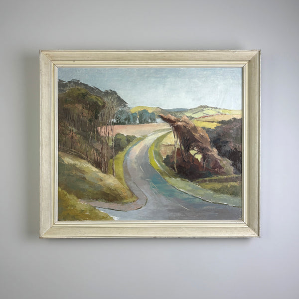 A vintage framed landscape oil painting hanging on a light gray wall. The painting depicts a winding road through rolling hills under a pale sky, enclosed in a distressed off-white wooden frame.