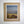 A vintage mid century framed 1964 dated and signed landscape painting on canvas, depicting a moody blue-gray sky over a golden field with dark reddish-brown shrubs in the foreground. The painting is matted with a beige linen mat and enclosed in a gold frame, hanging on a plain white wall.