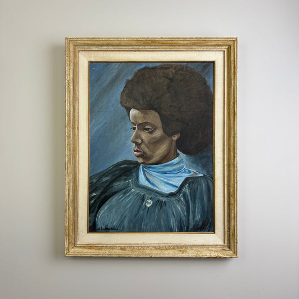 A MCM mid century modern framed portrait painting of a Black woman with an afro hairstyle, wearing a light blue collar and dark blue dress, set in a distressed gold frame and hanging on a light gray wall. VIntage art in good condition 