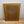 The back view of a square framed picture with a light brown wood frame and a brown cork backing, held together by a metal wire support, set against a plain light gray wall and wooden floor.
