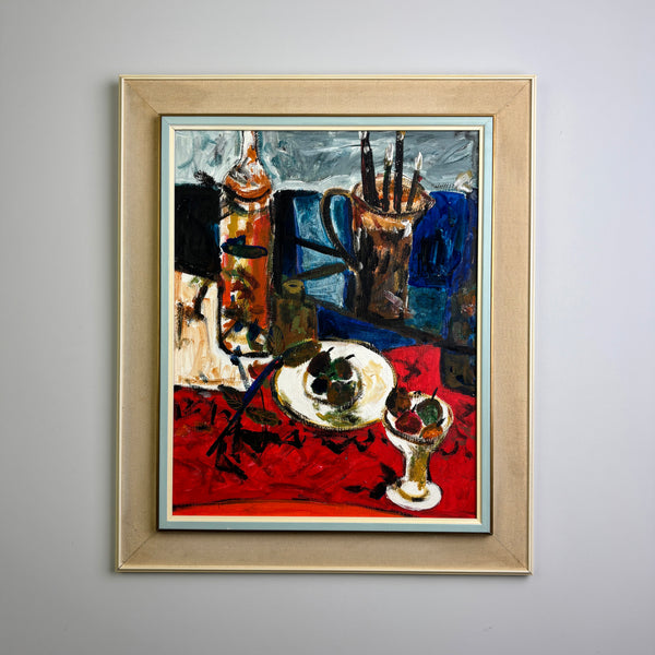 A framed vintage still life oil painting featuring a red tablecloth, a white plate with dark green olives, a small glass with red fruit, and a bottle, housed in a light wood frame and displayed on a neutral gray wall. good vintage condition with original vintage frame