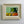 A framed mid century still life painting featuring a green teapot on a table with lemons and oranges, set in a distressed beige linen frame against a light gray wall.