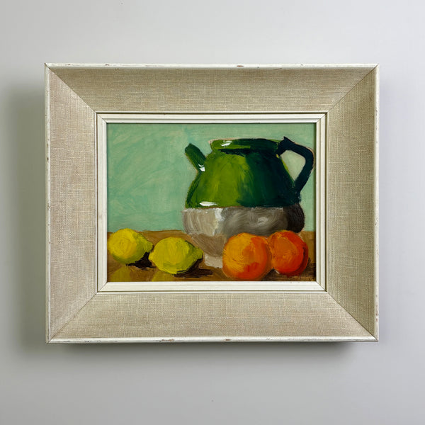 A framed mid century still life painting featuring a green teapot on a table with lemons and oranges, set in a distressed beige linen frame against a light gray wall.