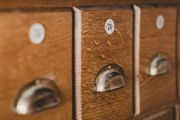 A close-up of a vintage wooden cabinet with three drawers, featuring brass semicircle pulls and small white round labels with handwritten numbers.